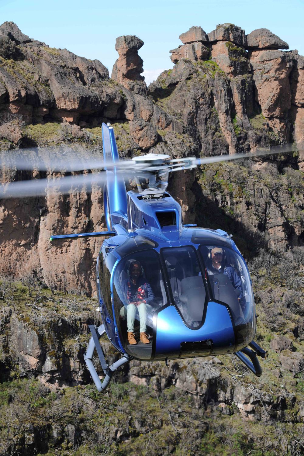 Passengers flying doors-open aboard a Tropic Air helicopter beside dramatic volcanic rock spires, pilot and guest visible in the cabin